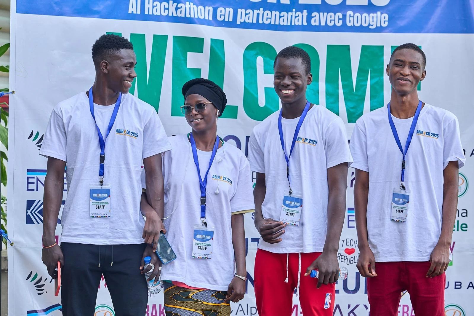 Quatre jeunes en t-shirt ABIHACK 2025, badge « Staff », sourient devant la bannière d’accueil.